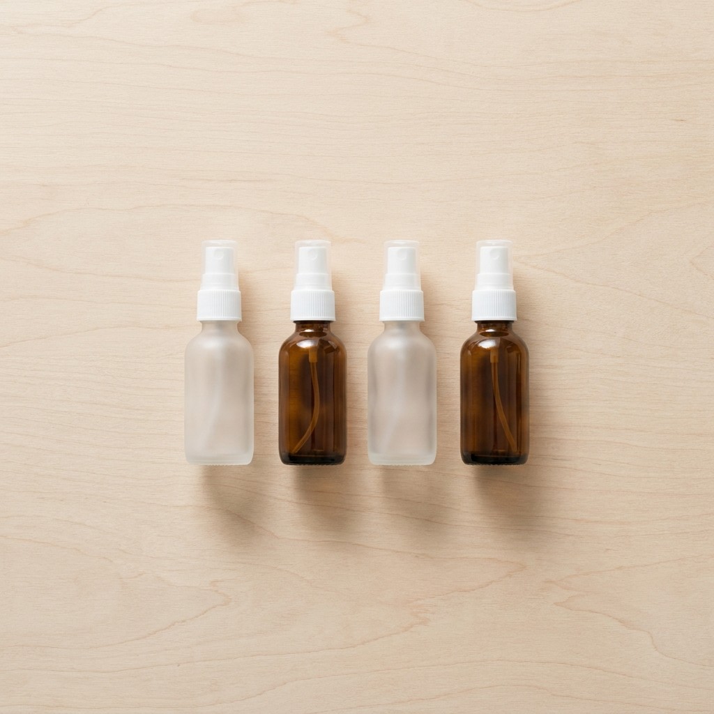 Four glass travel toiletry spray bottles in frosted and amber finishes on wood